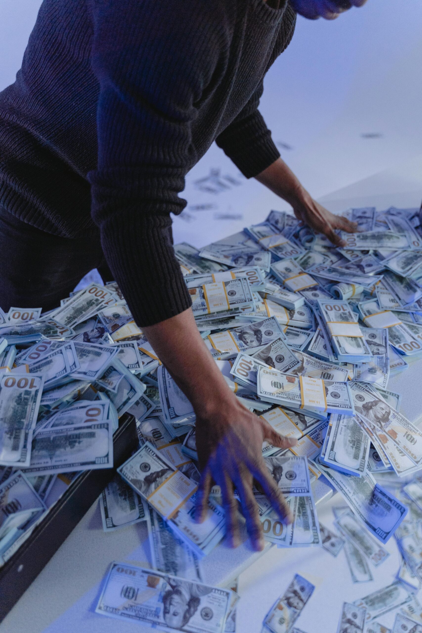 Adult man organizing a massive pile of US dollar bills indoors in dynamic lighting.